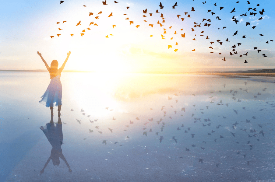 Una mujer en una playa al amanecer con una bandada de pájaros en el horizonte.