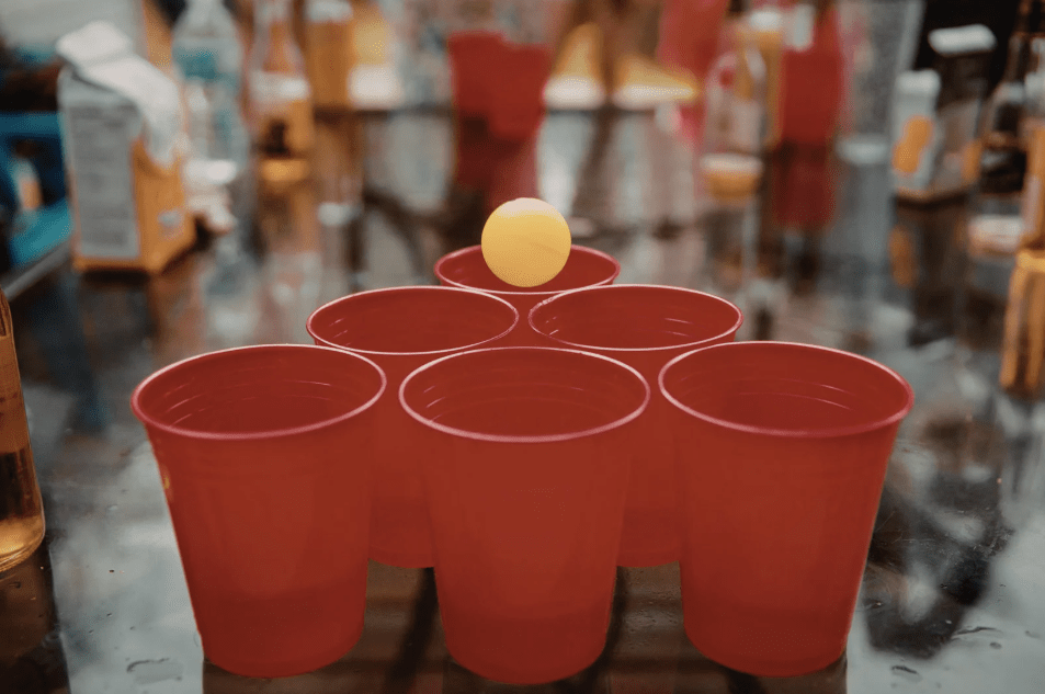 Seis vasos rojos puestos en triángulo, y una pelota de tenis entrando en uno de ellos.