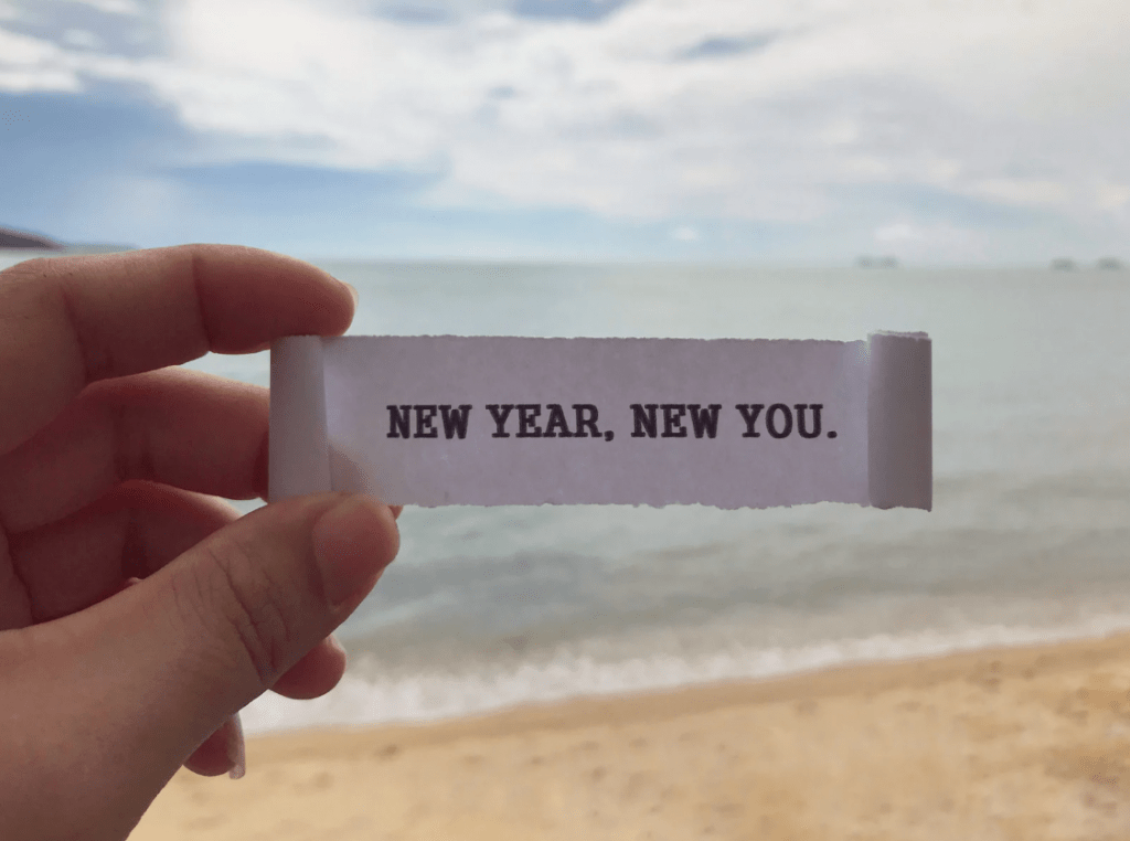 Unos dedos sujetando un cartel que pone "New year, new you" frente a la playa.