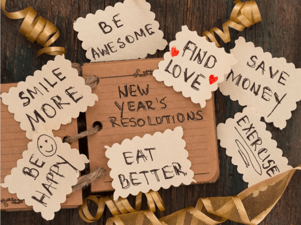 Un cuaderno que reza "New year's resolutions" y carteles alrededor que dicen "be awesome", "find love", "save money", "exercise", "eat better", "be happy", "smile more".