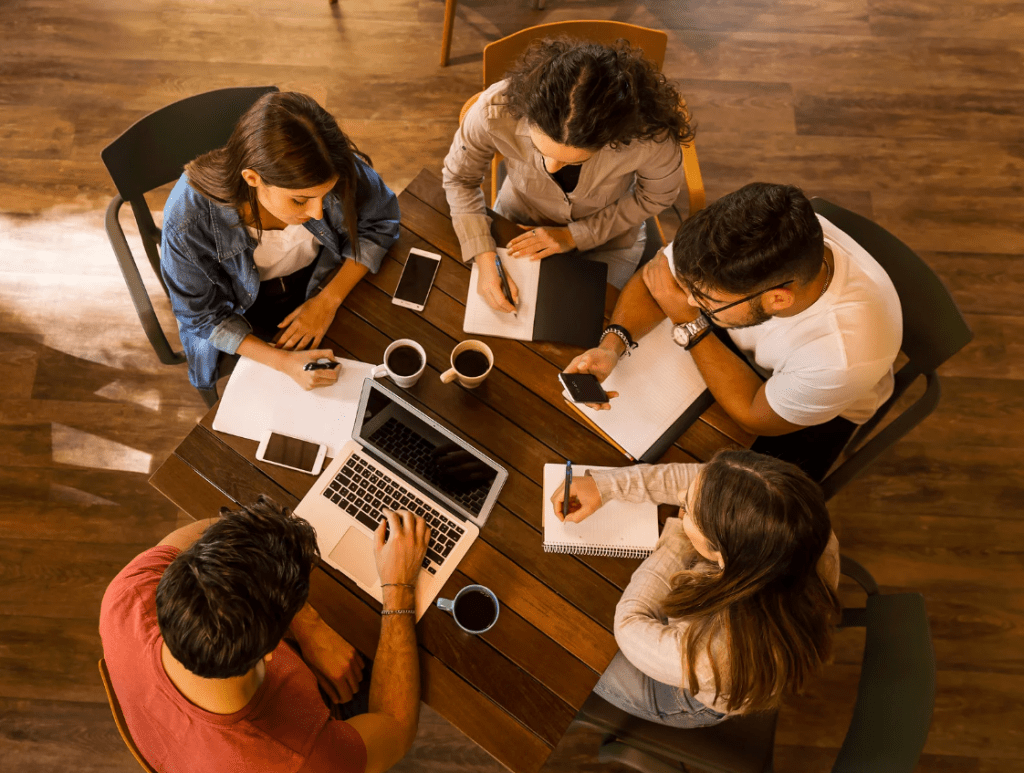 Cinco jóvenes en una mesa, trabajando con dispositivos electrónicos y cuadernos.