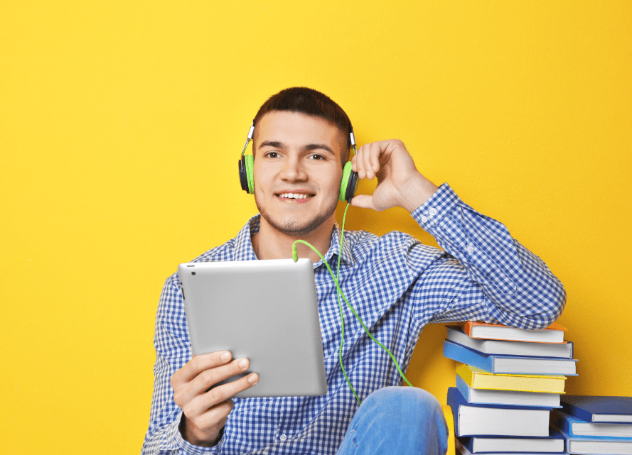 Un joven con cascos escuchando en su tablet, con una pila de libros al lado.
