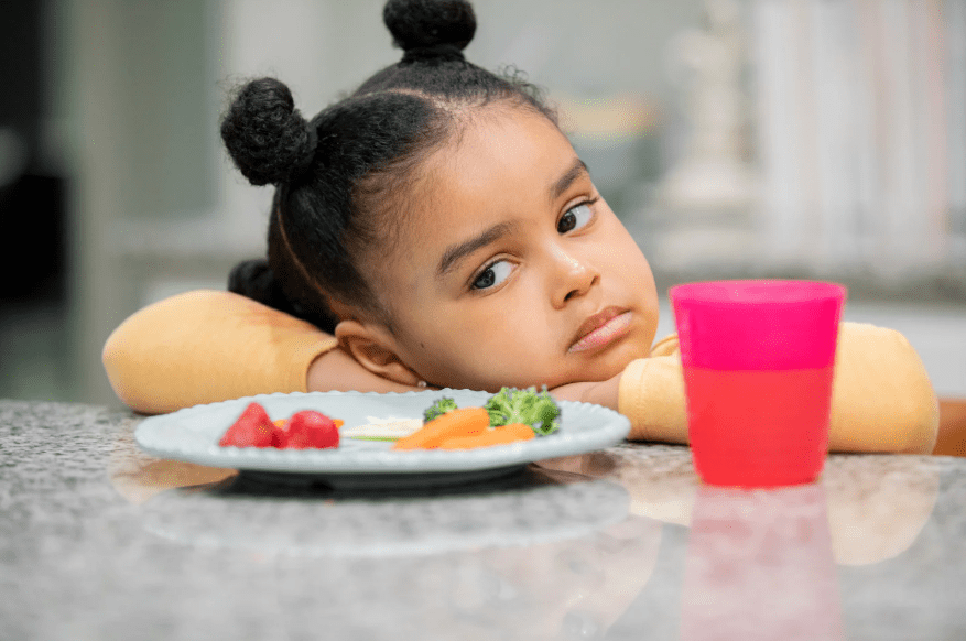 Una niña con cara de aburrimiento y un plato de verduras frente a ella.