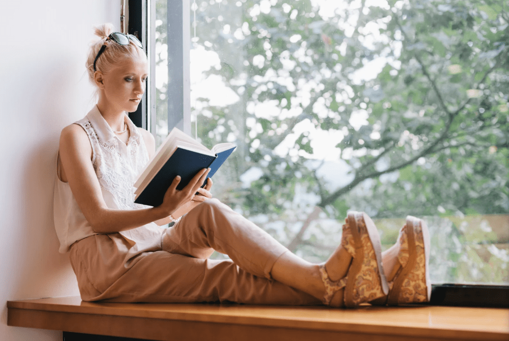 Una joven lee un libro sentada junto a una ventana.