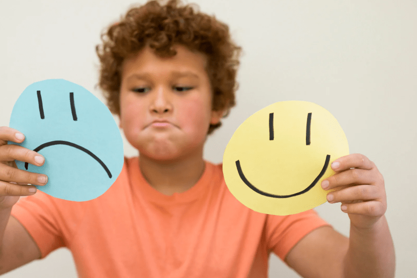 Un niño sujetando y mirando dos caras recortadas: una triste, y otra contenta.