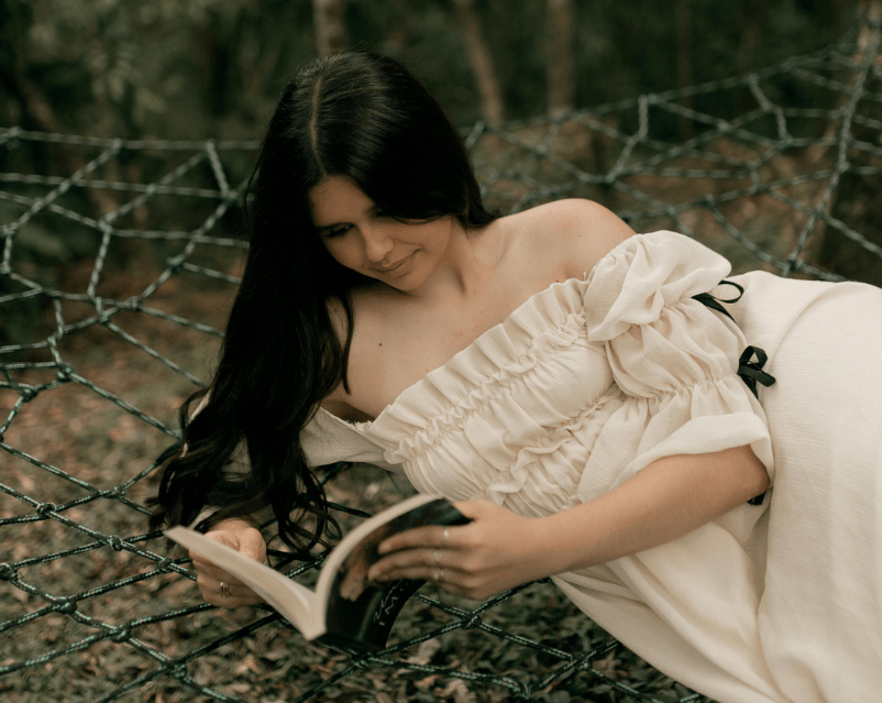 Una joven en un vestido blanco, tumbada en la naturaleza, lee un libro.